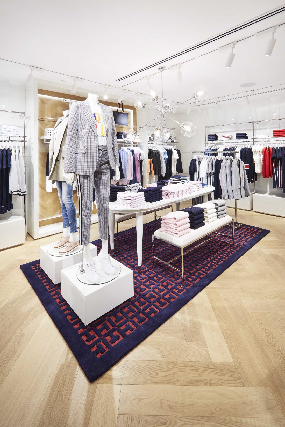 Fashion retail display with mannequins, folded knitwear and tailored garments arranged on patterned rug and timber flooring.