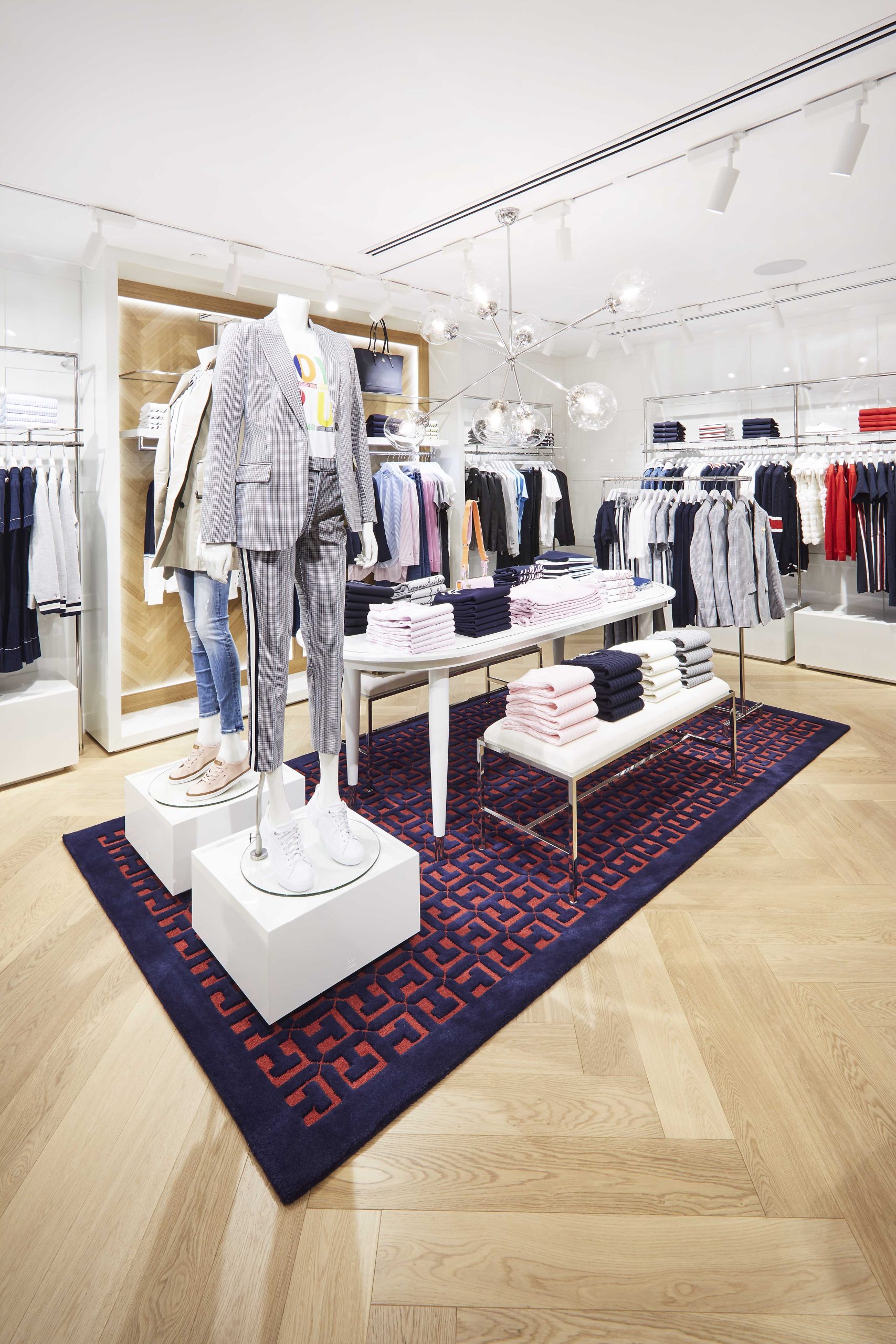 Fashion retail display with mannequins, folded knitwear and tailored garments arranged on patterned rug and timber flooring.