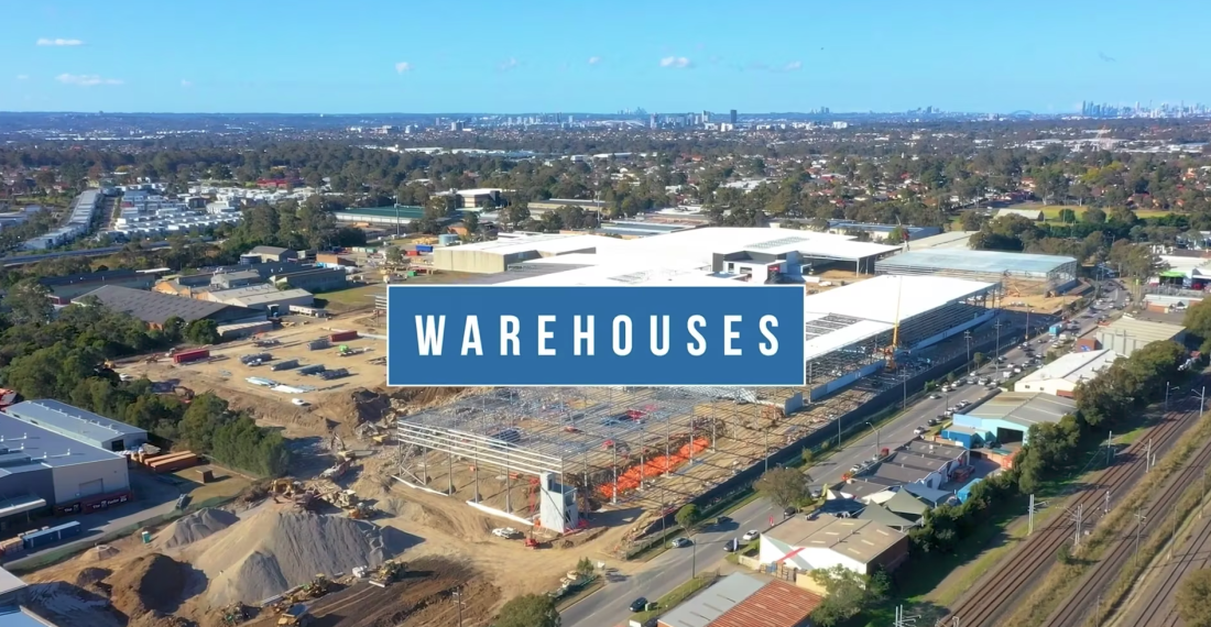 Aerial drone view of warehouse construction site with steel framing, surrounding industrial buildings and distant city skyline.