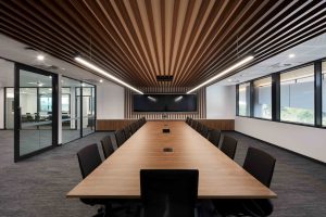 Contemporary boardroom with long timber table, black chairs, timber batten ceiling and dual wall‑mounted screens.
