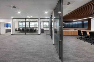 Modern open‑plan office with glass meeting rooms, large screen in timber‑lined boardroom and grey carpet throughout.