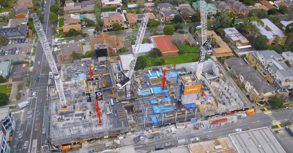 Aerial view of a large multi‑storey construction site with tower cranes and bright blue formwork surrounded by residential streets and houses in an inner‑suburban neighbourhood.