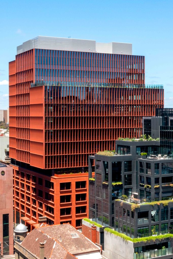 Aerial close-up of a completed red commercial tower with glazed facade and neighbouring green-roof buildings.