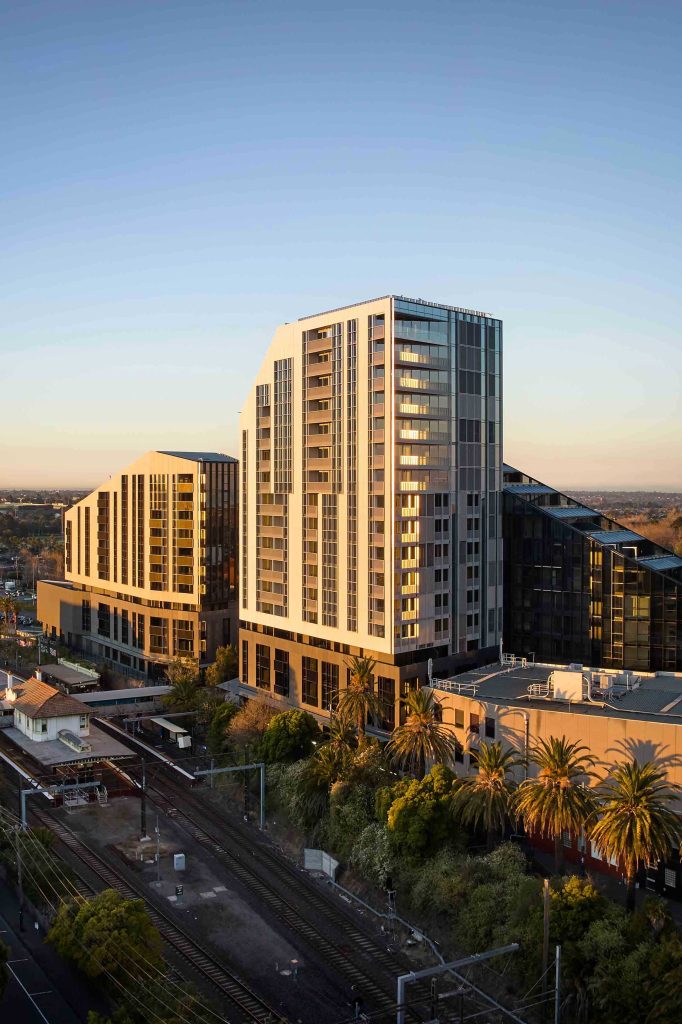 Golden hour aerial view of stepped residential buildings above a rail corridor lined with palm trees.