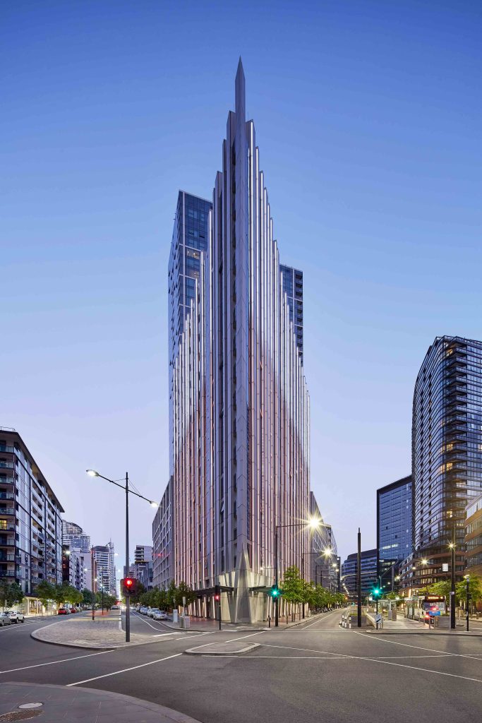 Evening street view of a slender glass tower forming a sharp prow at a city intersection.