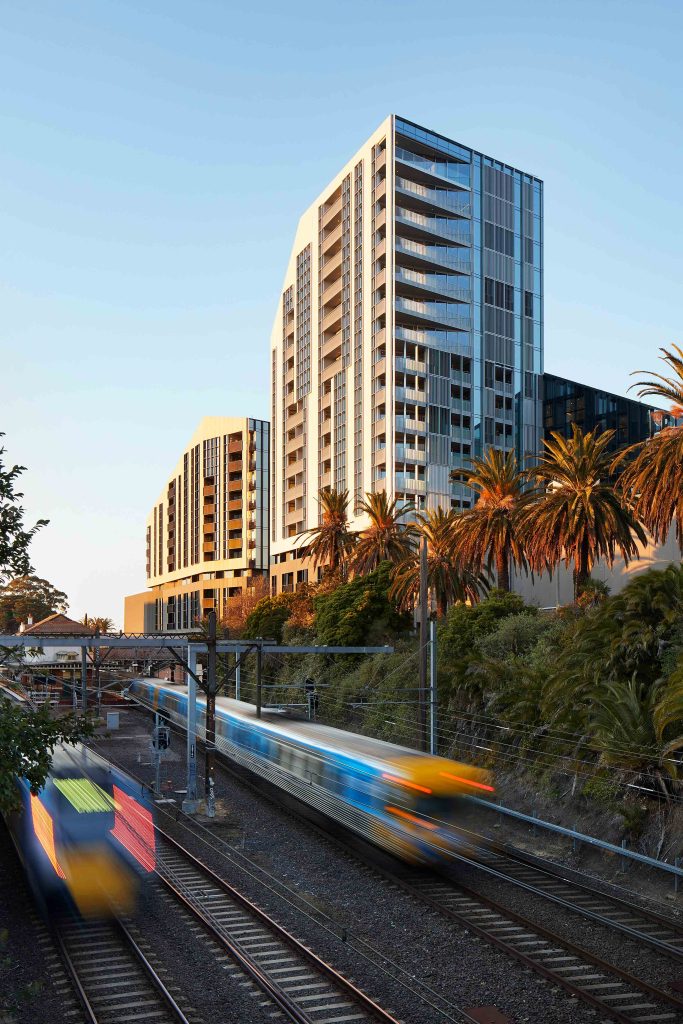 Residential towers above a rail corridor with blurred trains passing through a lush, palm-lined cutting.