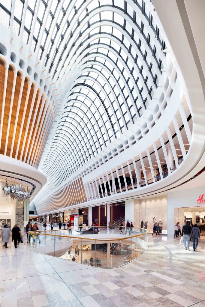 Soaring white retail atrium with curved bays and glass balustrades overlooking shoppers on multiple levels.