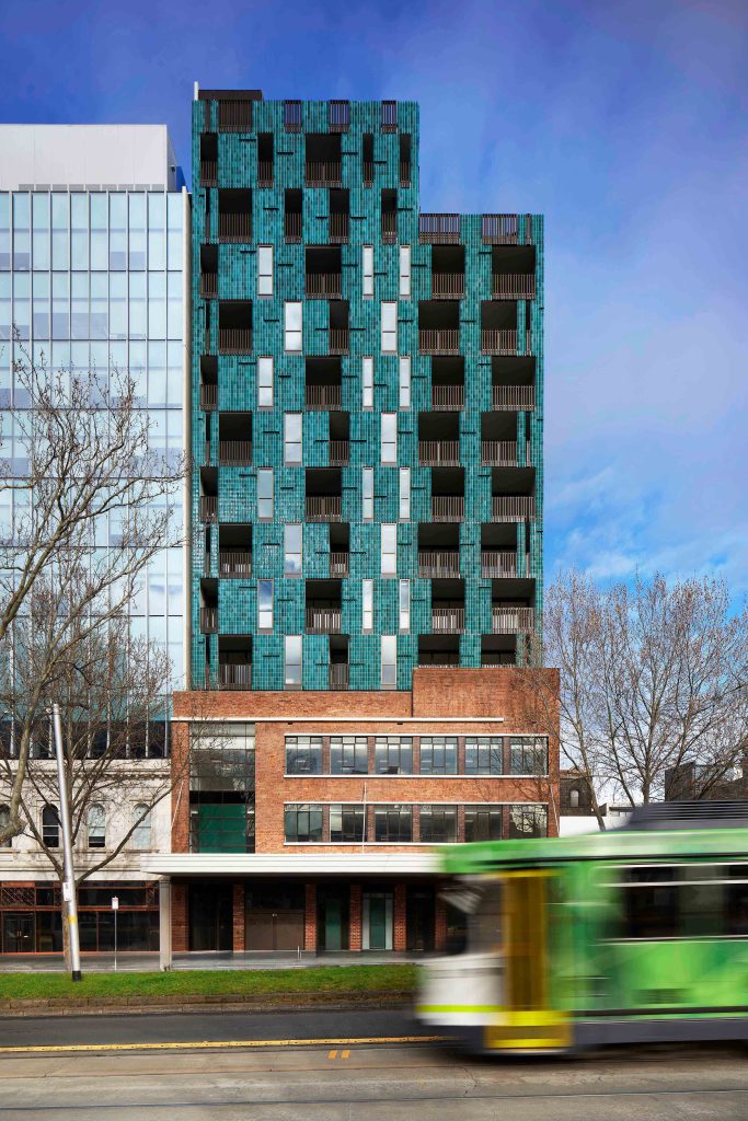 Teal-green residential tower with stepped form and chequered facade rising above heritage brick podium and tram.