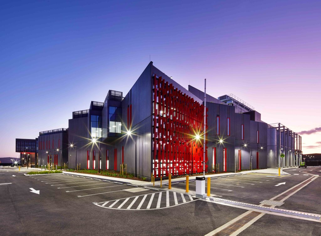 Industrial building with dark corrugated cladding and red perforated screen panels glowing at twilight in car park.