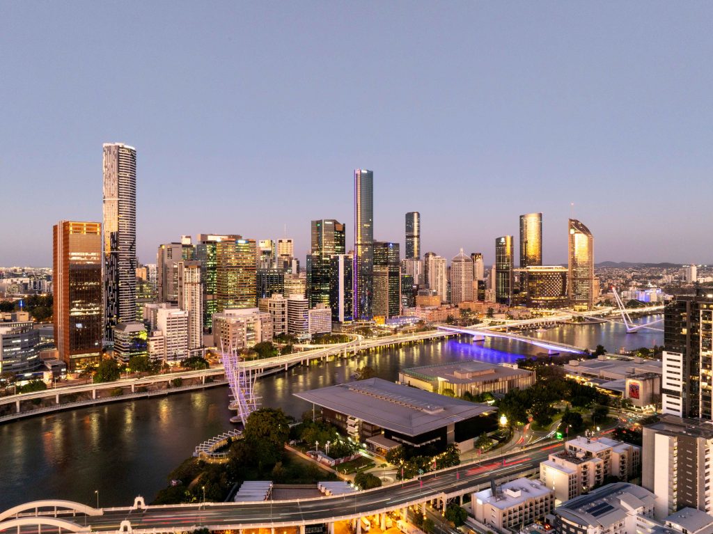 Evening Brisbane skyline with towers and river reflecting warm light under purple sky at dusk.