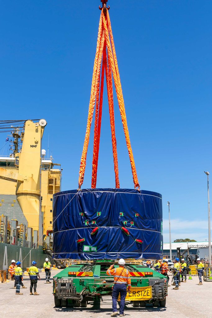 Large wrapped load suspended from orange slings as workers manage an oversized lift at a port.