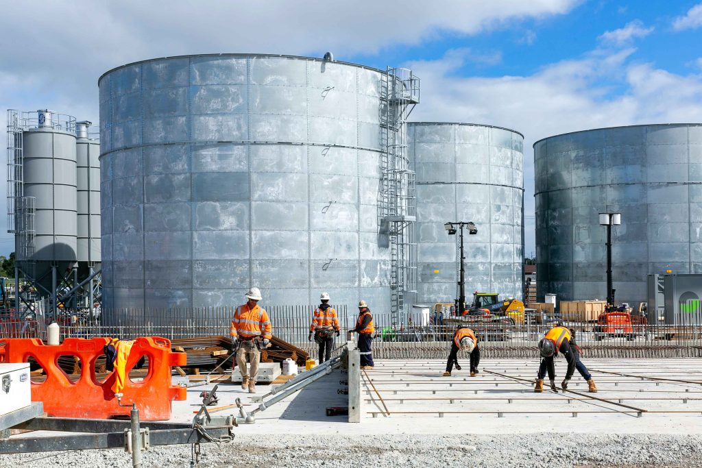 Construction workers installing reinforcement on a slab in front of large cylindrical storage tanks.