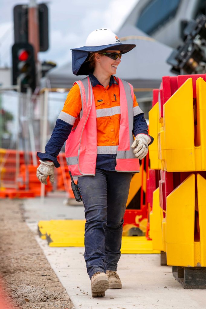 Construction supervisor in pink vest walking beside yellow and red barriers on an urban work site.