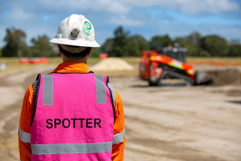 Spotter in pink vest watching an orange skid steer operating on an open civil site.