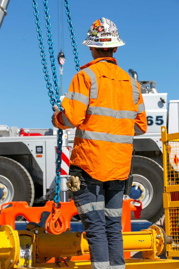 Rigger in orange PPE holding blue chains beside heavy lifting equipment on a sunny site.