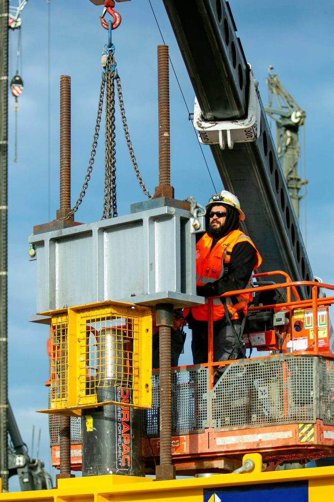 Worker on elevated platform managing chains connected to a large steel assembly beneath a crane boom.