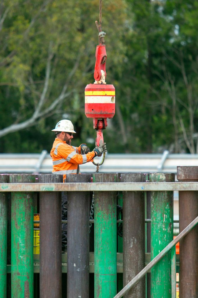 Construction worker in orange PPE guiding a red crane hook above sheet piles on site.