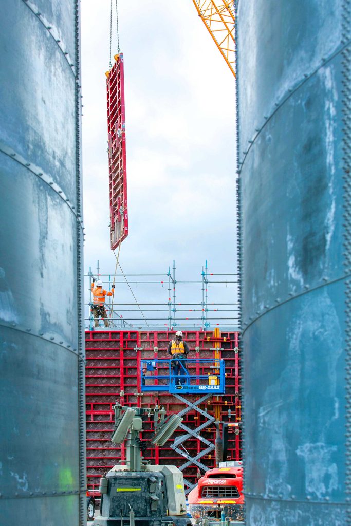Workers guide a tall red formwork panel being lifted between two large steel tanks.
