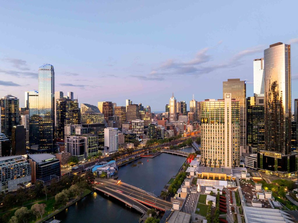 Dusk aerial view of Melbourne Southbank skyline with towers reflecting across the Yarra River and pedestrian bridge.