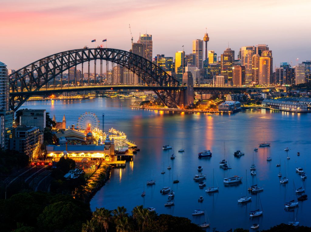 Sydney Harbour at dusk with Harbour Bridge, CBD towers and moored yachts reflecting soft pink light.