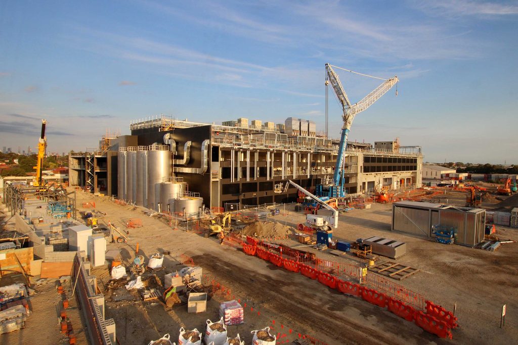 Wide construction site view showing an industrial facility rising with cranes, silos, and services at sunset.