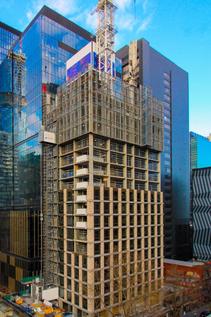 High-rise tower mid-construction with exposed structure and facade panels rising among finished CBD buildings.
