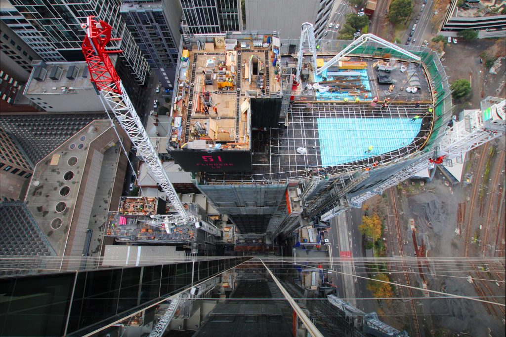 Dramatic top-down view of a high-rise construction deck with tower cranes above city streets and rail lines.