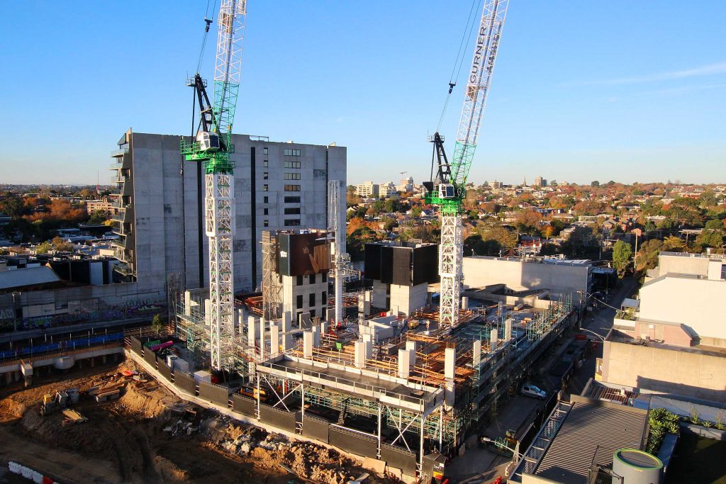 Elevated view of a multi-storey structure rising with twin tower cranes above surrounding suburban rooftops.