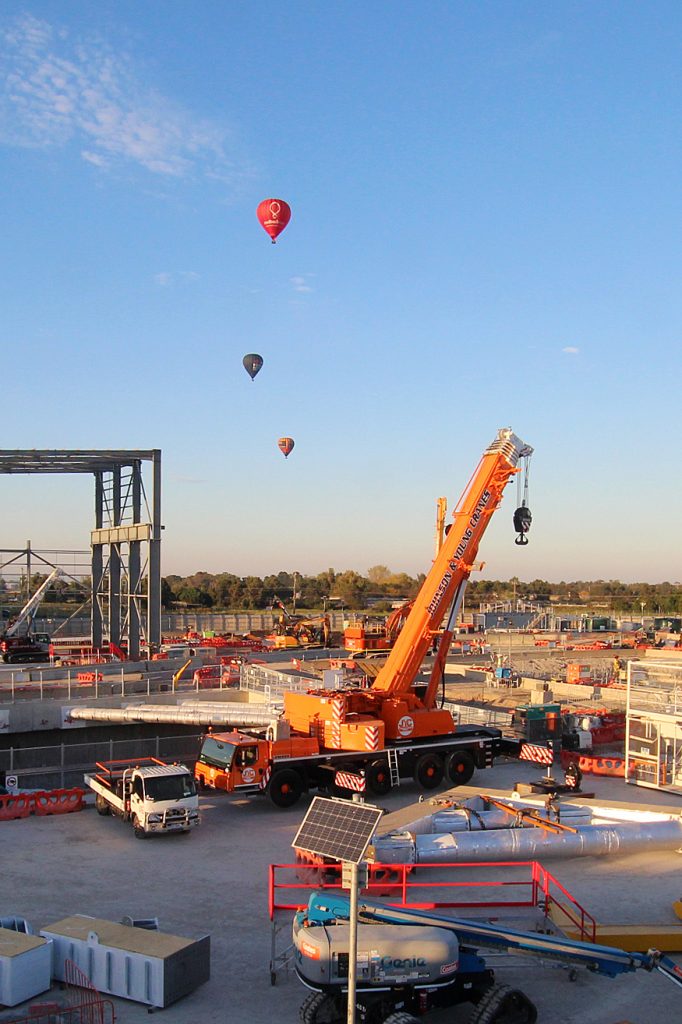 Mobile crane on an early-morning construction site with hot air balloons rising in the sky behind.
