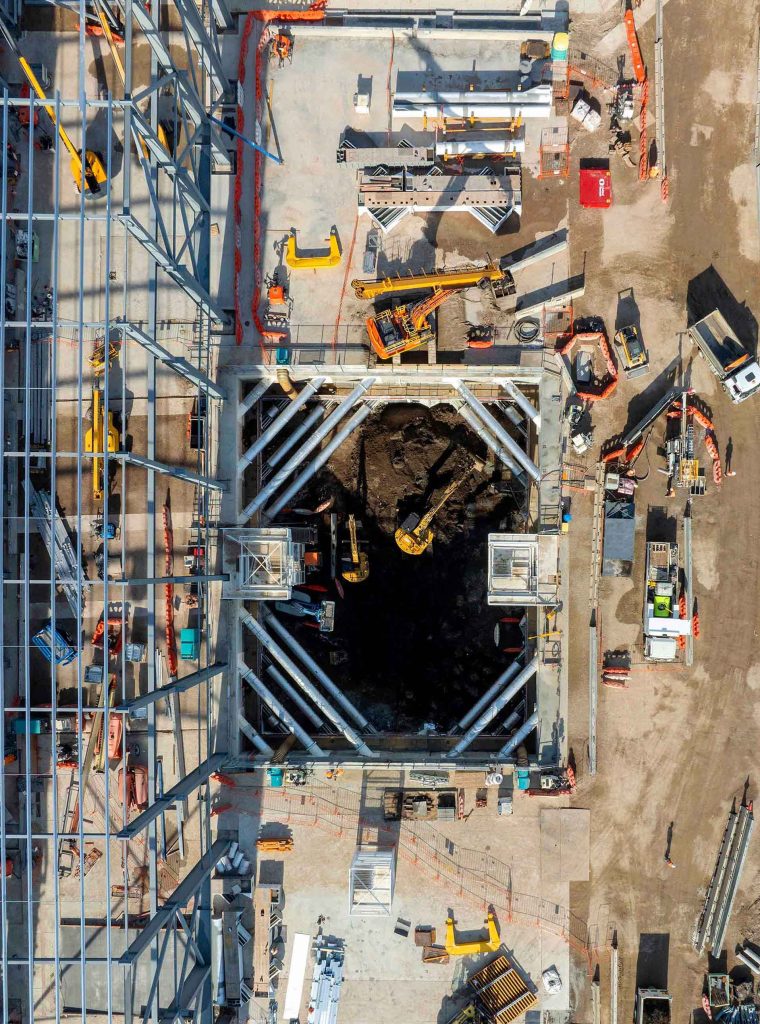Aerial view of deep excavation framed by braced retaining walls and surrounded by construction plant and materials.
