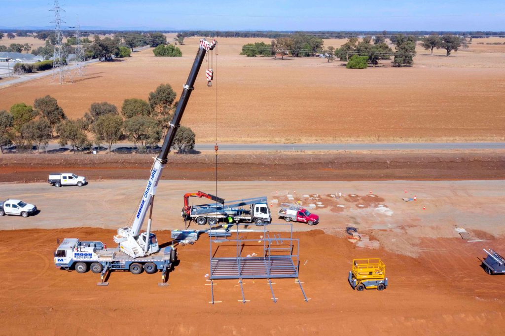 Aerial view of mobile crane and trucks assembling a steel frame structure on a rural construction site.