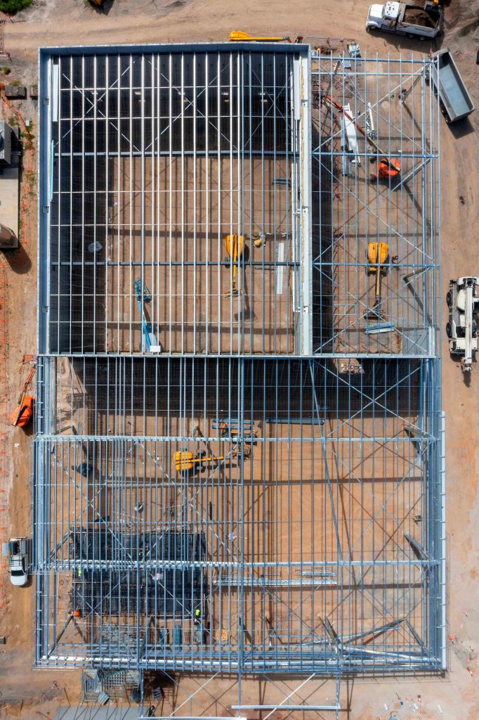 Aerial view looking down on a large steel-framed industrial building under construction with access equipment inside.