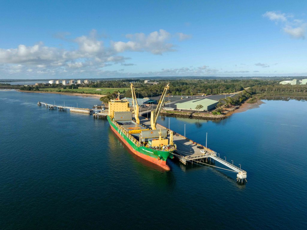 Aerial view of a large cargo vessel berthed at an industrial wharf with warehouses and coastline in the background.