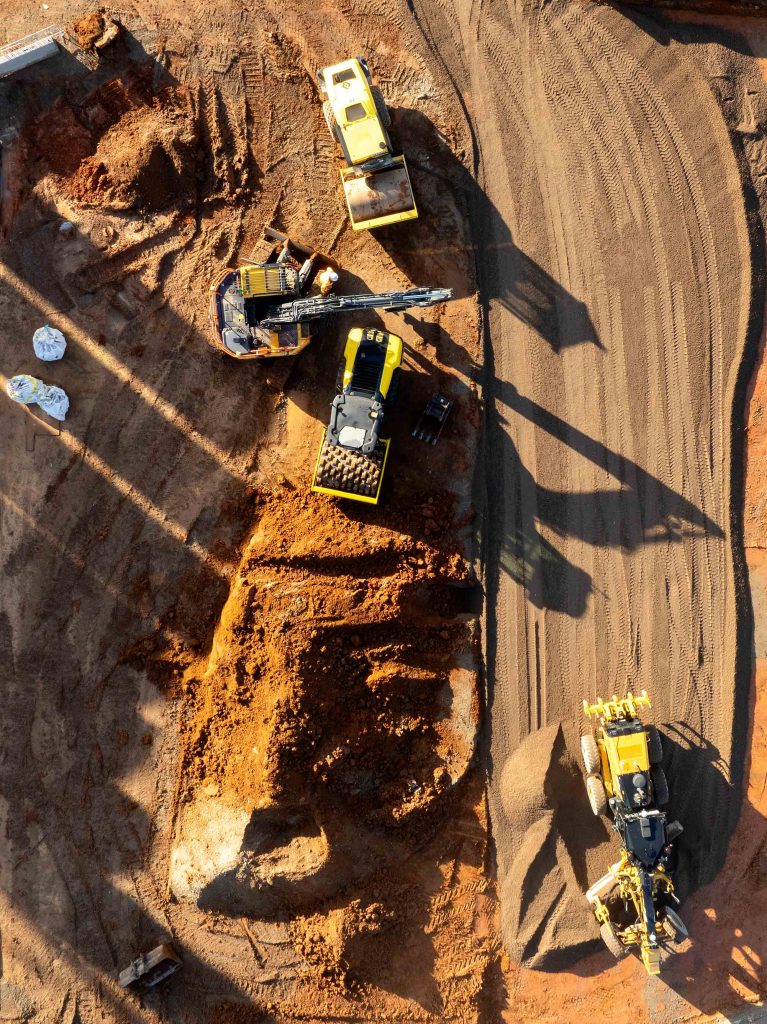 Aerial view of excavator, compactor and grader shaping and compacting earthworks on a construction site.