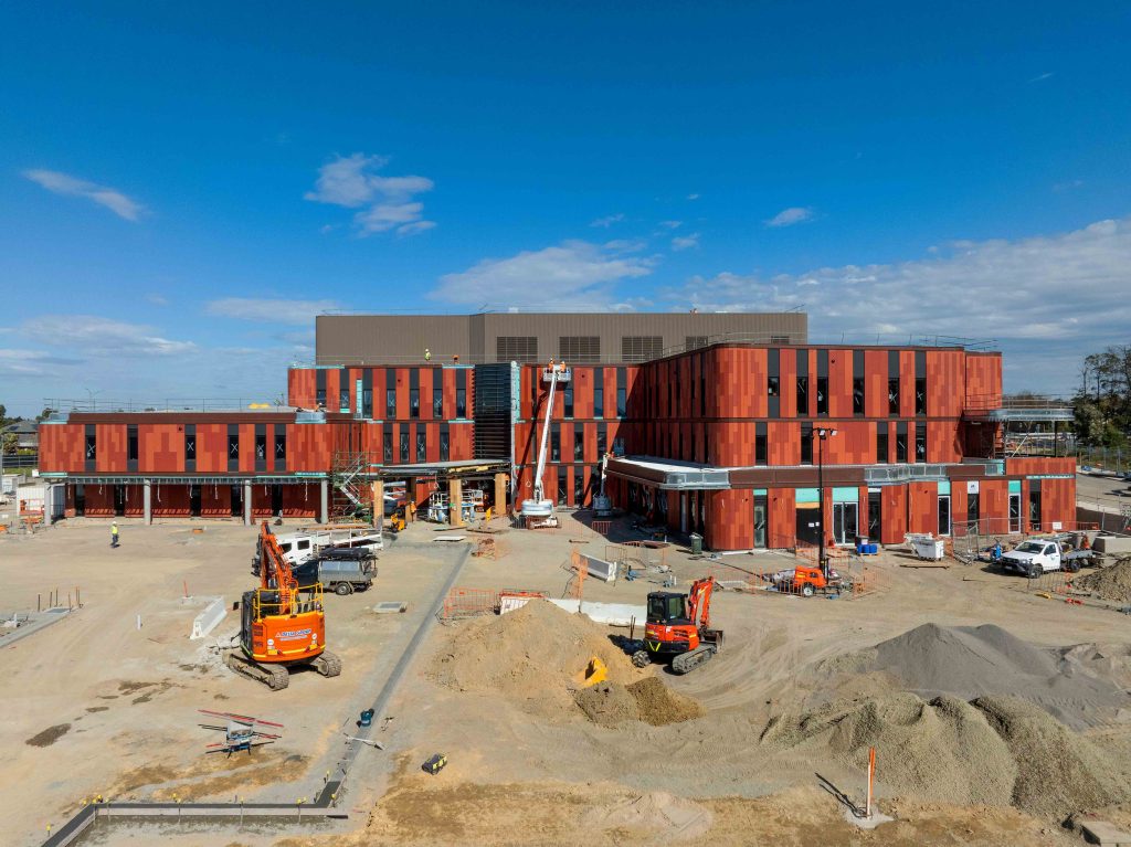 Aerial view of a partially completed red-clad building with earthworks and construction plant in the foreground.