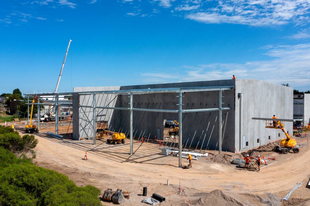 Aerial view of precast concrete warehouse walls with steel frame installation and workers on boom lifts.