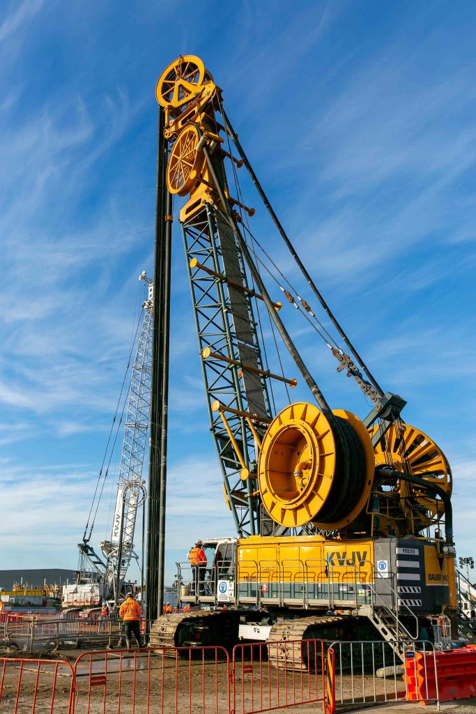 Large Bauer crawler drilling rig with tall mast, cable reels and workers in high-visibility clothing operating at a civil construction site.