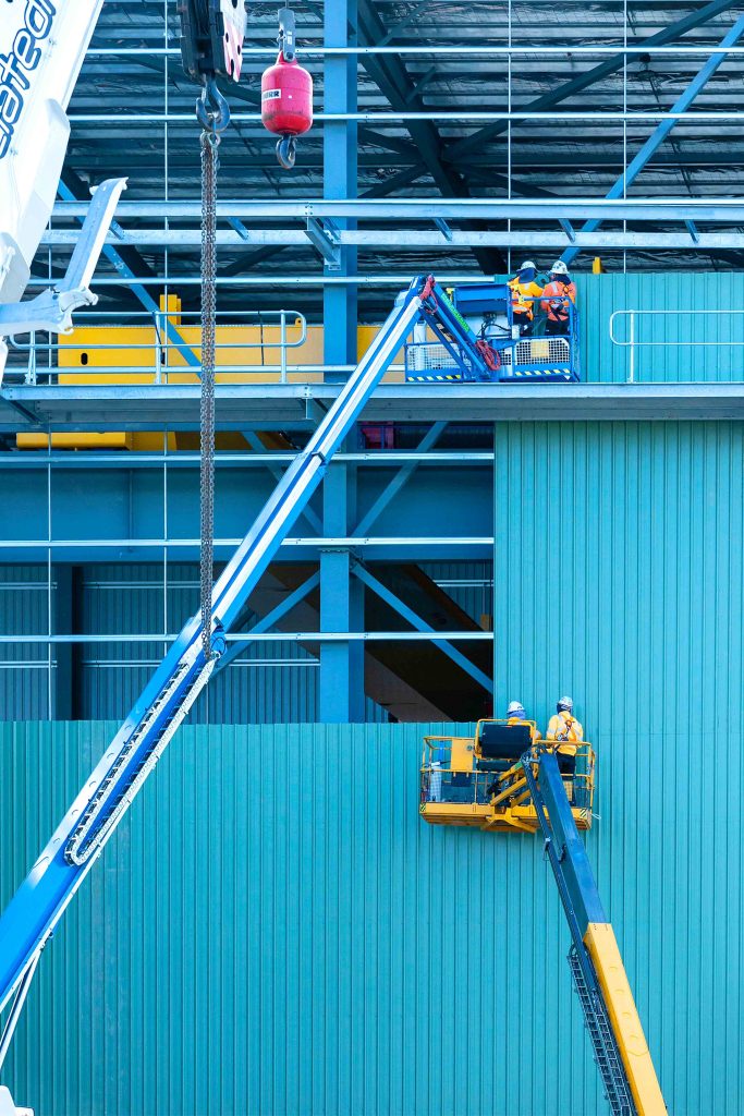 Construction workers in elevated work platforms installing façade cladding on a steel-framed industrial building, with crane hook in foreground.