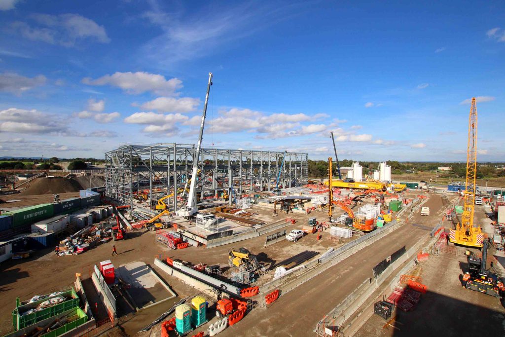 Elevated wide view of large industrial construction site with steel frame structure, multiple mobile cranes, excavators, materials and temporary site facilities.