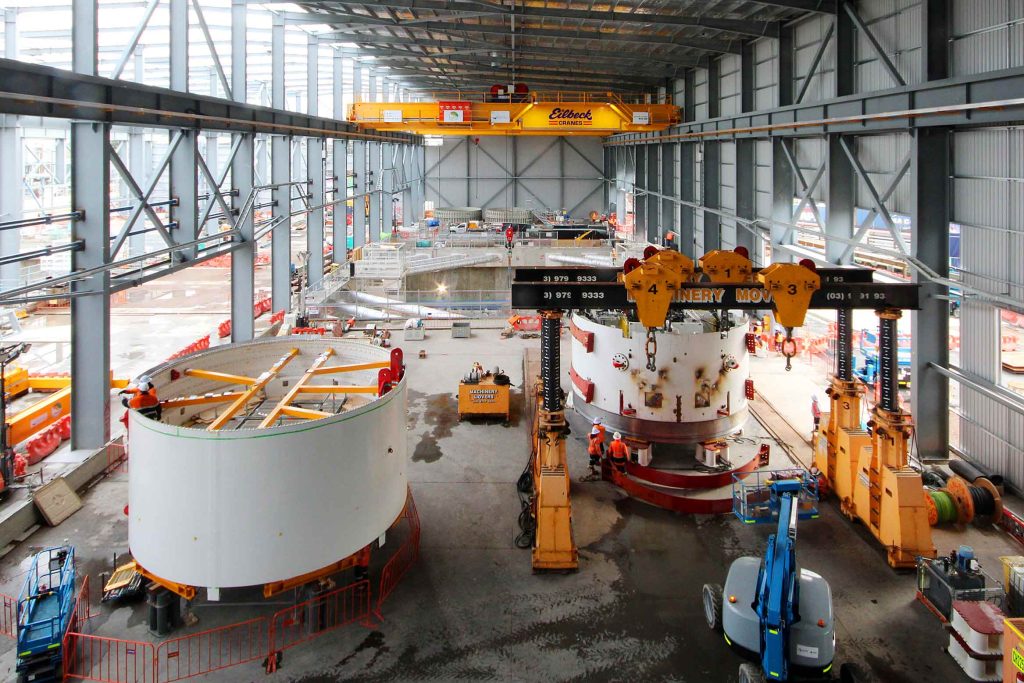 Heavy industrial construction hall with overhead crane, mobile gantry system and workers assembling large cylindrical steel components.