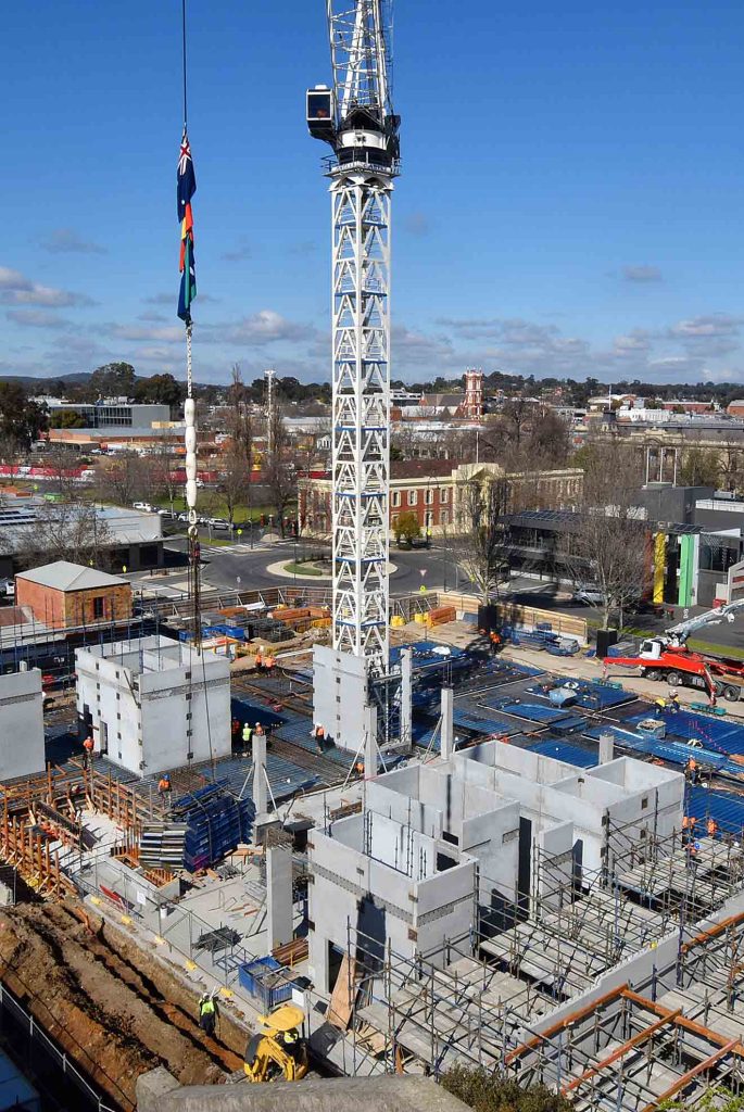Elevated view of a construction site with tower crane lifting a flag-wrapped load above precast concrete walls, formwork and workers.