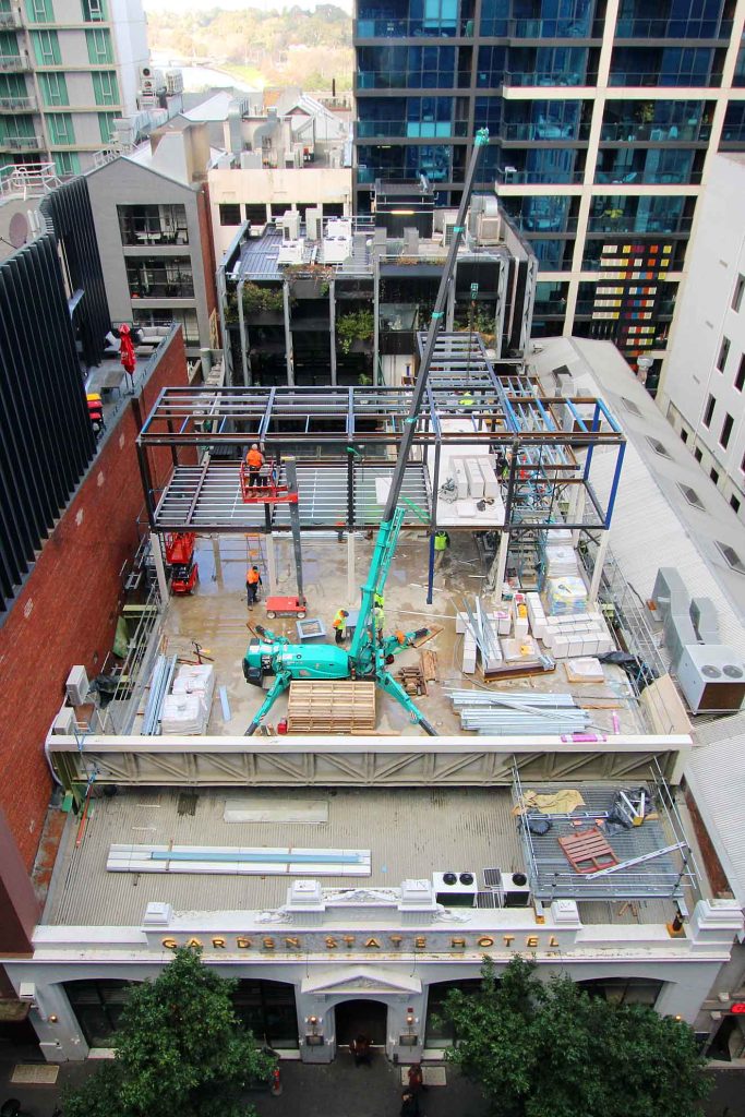 High-angle view of a compact urban construction site with steel frame, mobile crane, elevated work platforms and workers above a heritage hotel façade.