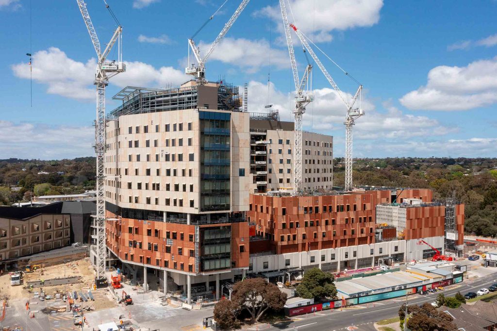Image 64 Alt text: Aerial view of a large multi-storey health or institutional complex under construction, with four tower cranes, articulated façades and active street-level works.