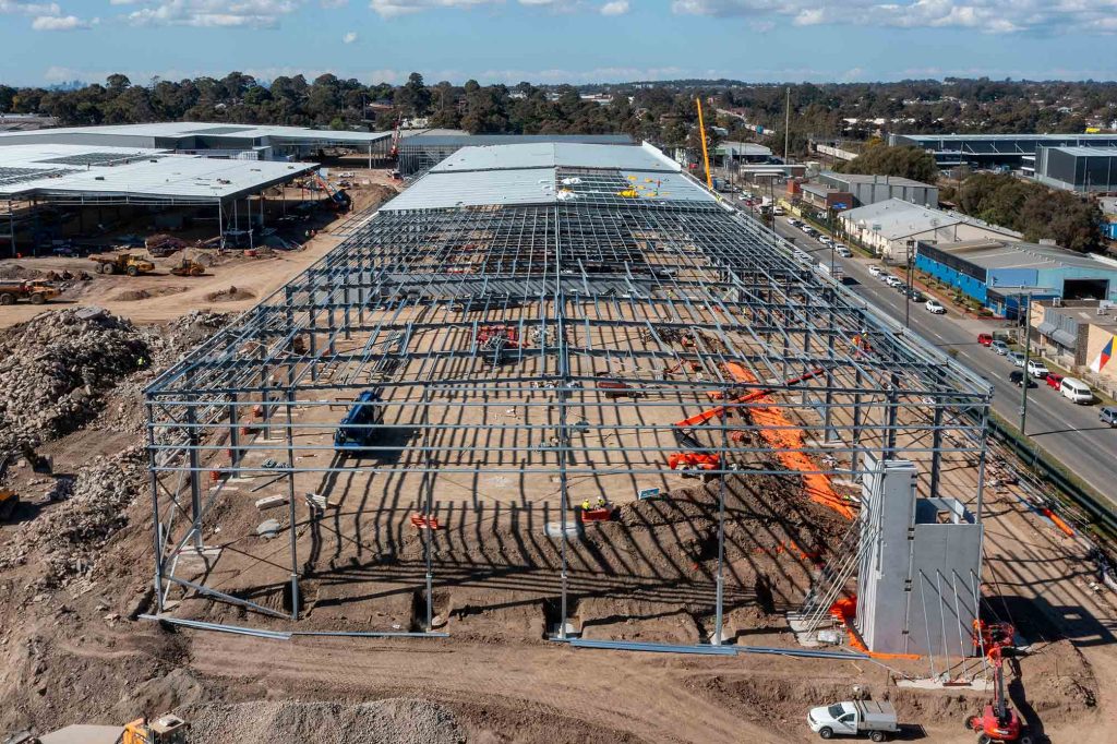 Image 65 Alt text: Drone view of a large steel-framed industrial building under construction, with roof purlins, earthworks plant and active works beside a busy road.