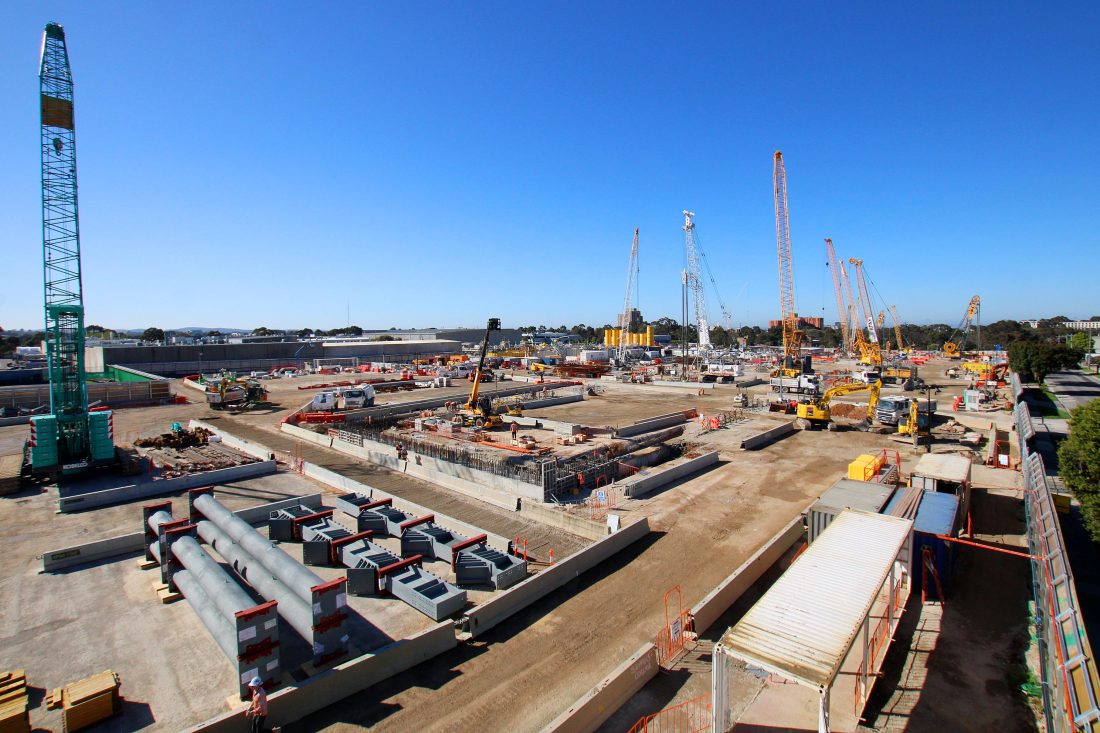 Elevated view of a large construction site with multiple cranes, concrete foundations, reinforcement, excavators and steel components stored in the foreground.