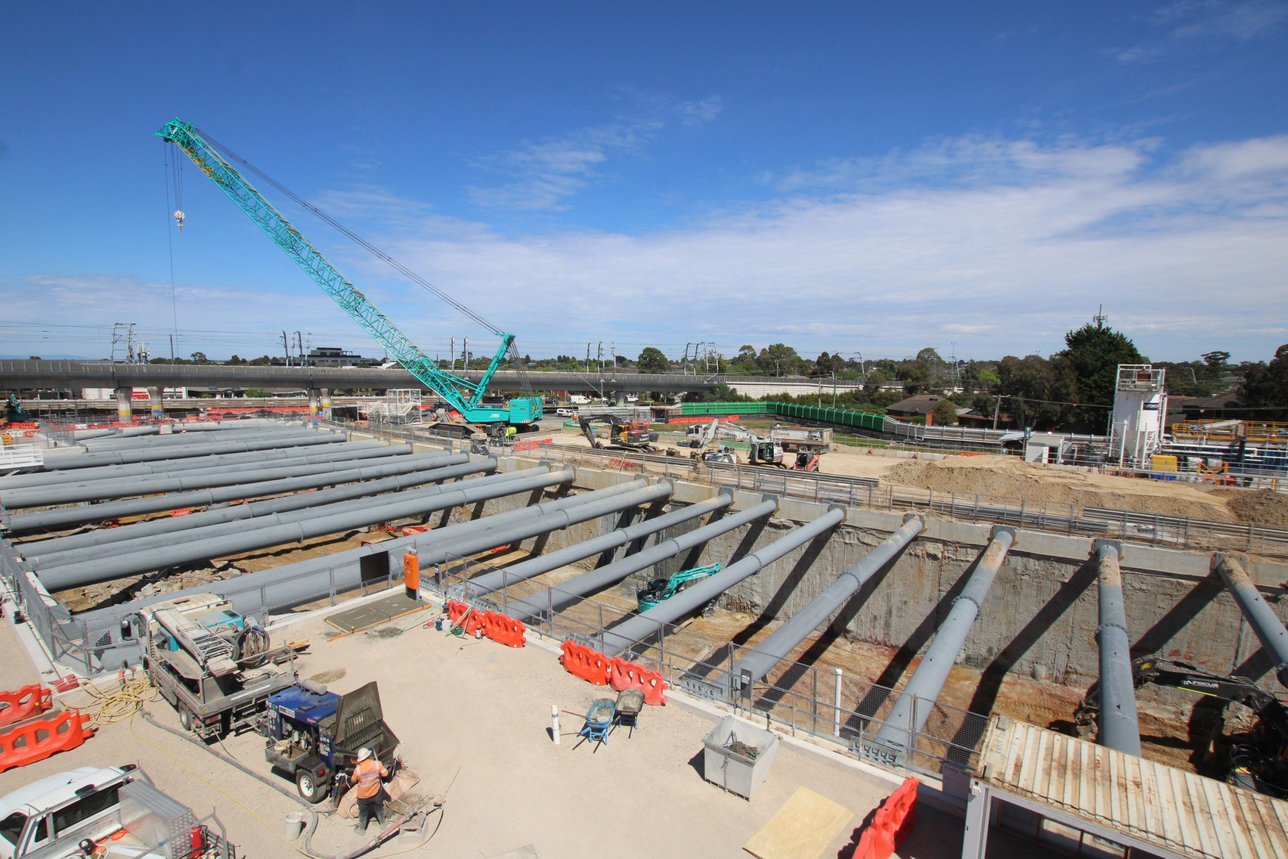Elevated view of a deep excavation crossed by rows of grey steel struts, with a turquoise crawler crane, excavators and an elevated railway behind.