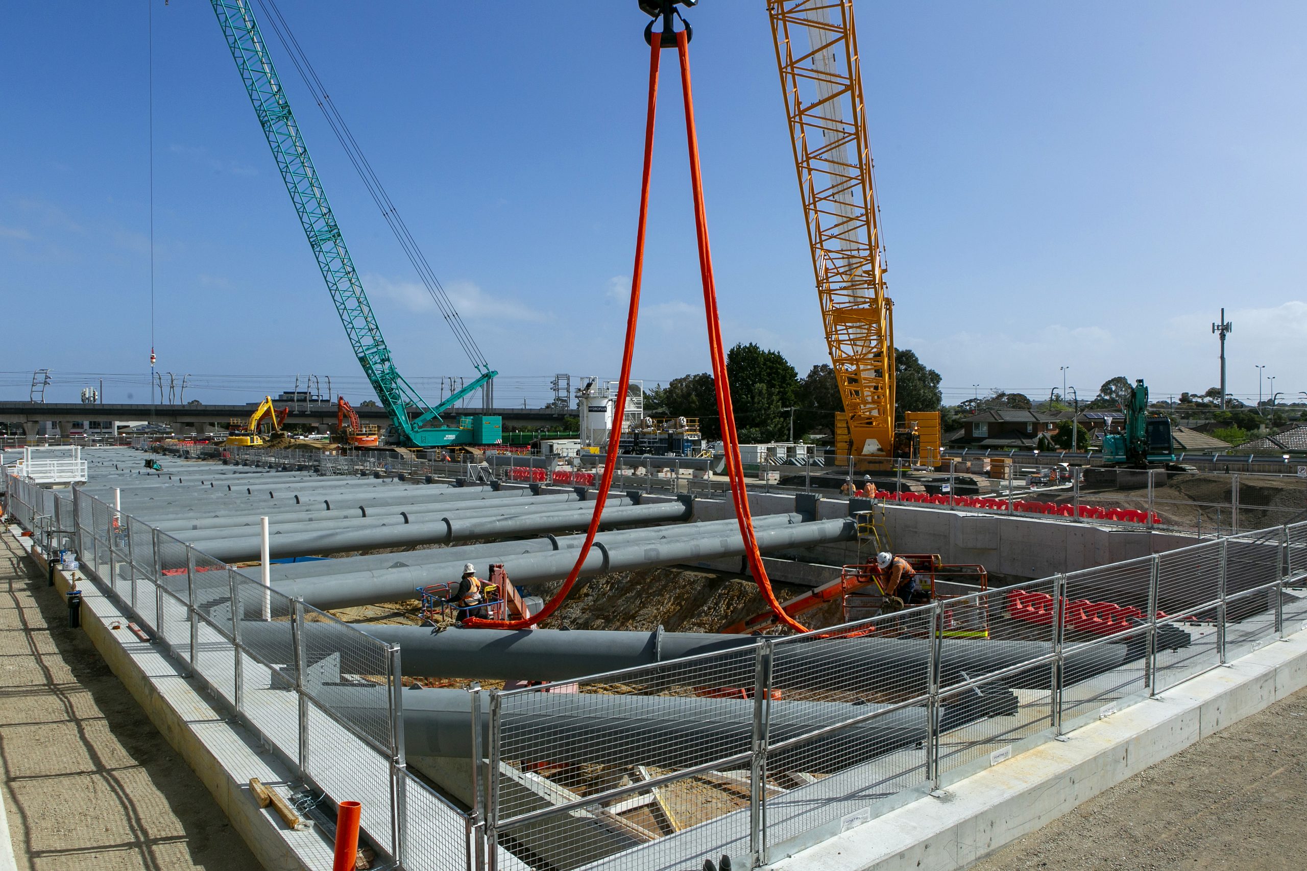 Crane lifting a large steel strut with orange slings above a braced excavation, with workers in boom lifts and crawler cranes behind.