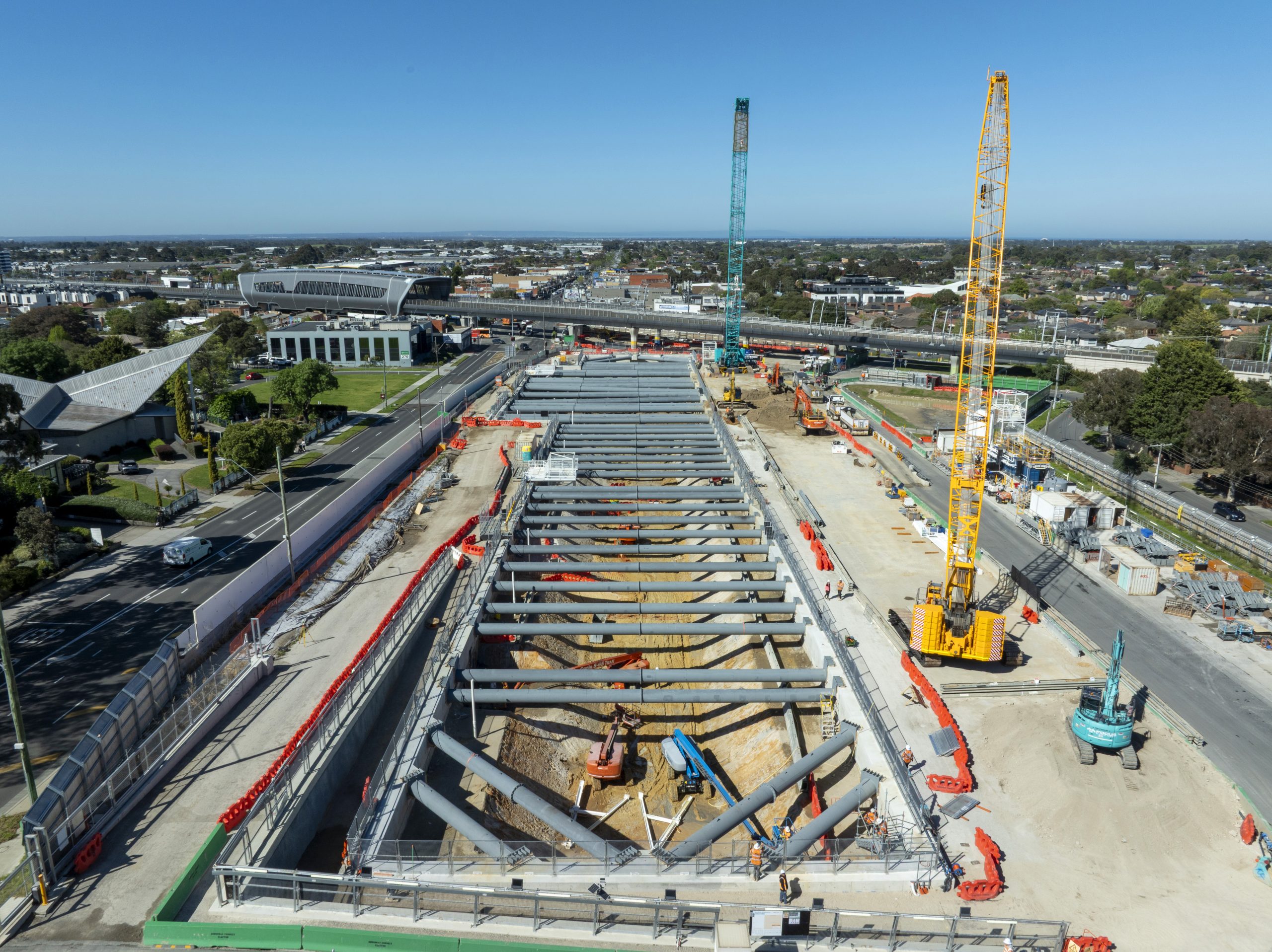 Aerial view of a deep rectangular excavation spanned by rows of steel struts, with crawler cranes, excavators and an elevated railway beyond. Construction visual documentation.